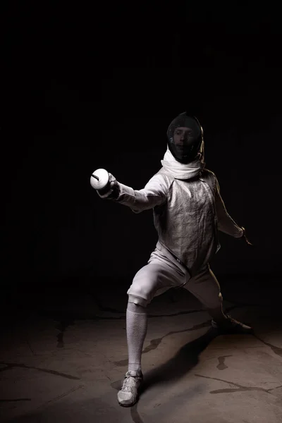 Portrait of male fencer in mask and foil performing fight in studio, dark key