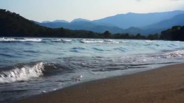 Arka planda dağlar ile kumlu bir plajda deniz dalgaları.