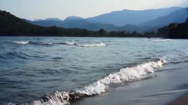 Arka planda dağlar ile kumlu bir plajda deniz dalgaları.