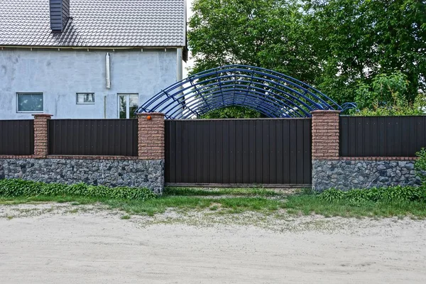 part of a long brown fence and gate of iron and stone in the street near the road from sand