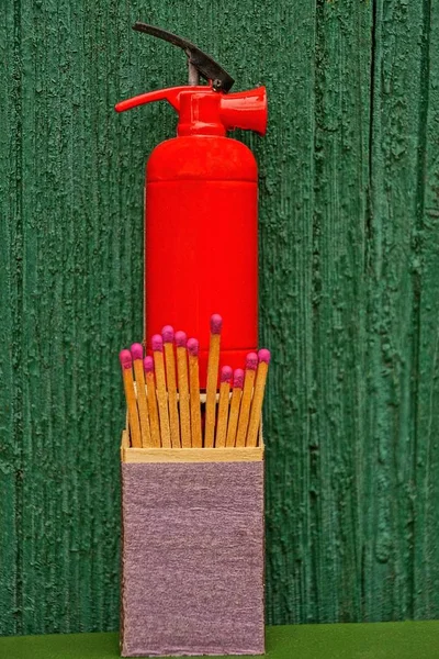 A small fire extinguisher stands on a box of matches near a green wooden wall