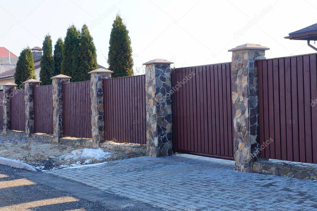 part of a long brown fence with a gate made of wooden planks and stones on the street along the gray asphalt road
