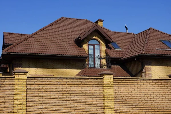 空に対するフェンスの後ろのタイル張りの屋根の下にバルコニーを持つレンガの茶色の家の一部 — ストック写真