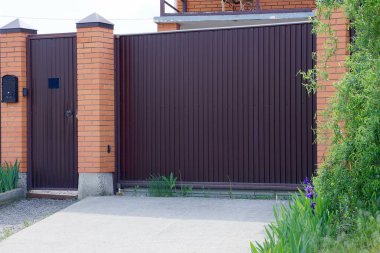 büyük bir kahverengi kapı ve yeşil bitki örtüsü dışında metal ve tuğla yapılmış bir çit parçası
