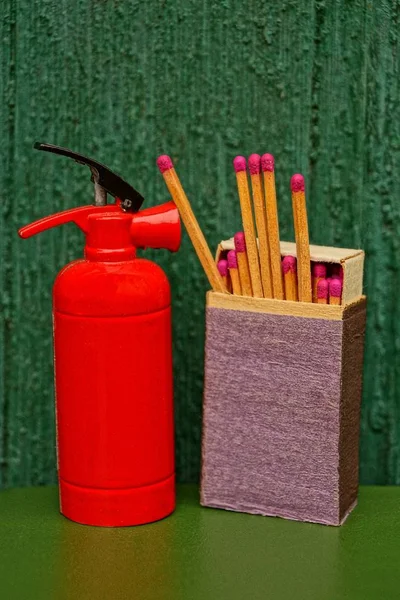 A small fire extinguisher stands on a box of matches near a green wooden wall