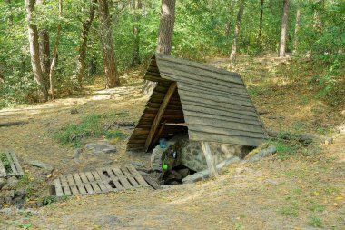 Ormandaki bir açıklıkta, gri ahşap çatının altında bir pınar olan pompa odası.
