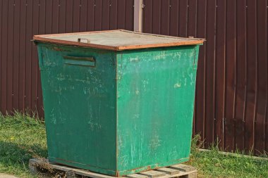 Bir yeşil metalimsi çöp tenekesi kahverengi bir çitin yanında.
