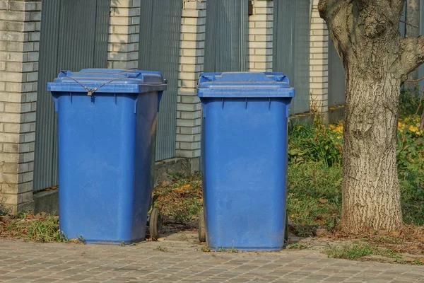 two blue plastic garbage cans on the street near a gray fence