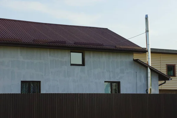 private gray house with windows under a tiled roof against a sky behind a fence