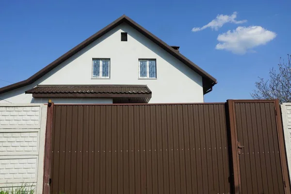 Kahverengi metal kapı ve mavi gökyüzüne karşı beyaz bir evin ön cephesinde beton bir çitin parçası.