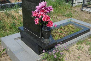 Siyah mermer bir vazo ve bir buket kırmızı yapay çiçek mezarlıktaki taş bir levhanın üzerinde duruyor.