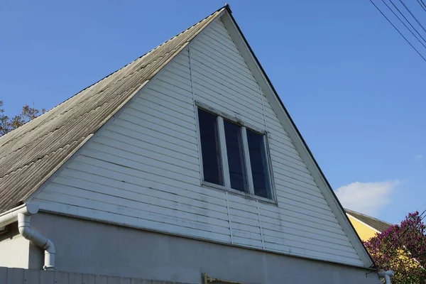 青い空に対して灰色のスレート屋根の下に窓のある田舎の家の白い木製の屋根裏部屋 — ストック写真