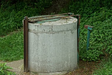 Bir eski gri kapalı beton kuyu yeşil bitki örtüsünün içinde caddede duruyor.