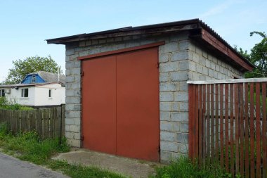 Kırsal bir sokakta kırmızı demir kapıları ve ahşap çitleri olan gri tuğlalı bir garajın ön cephesi.