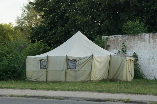 one large green military tent made of tarpaulin with windows stands in the grass by a gray road