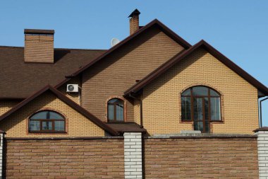 Çitin arkasındaki mavi gökyüzüne bakan kahverengi tuğlalı evin tavan arası.