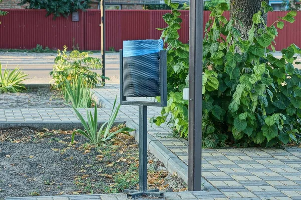 公園内の灰色の歩道に青い袋のついた黒い金属のゴミ箱が1つあります — ストック写真