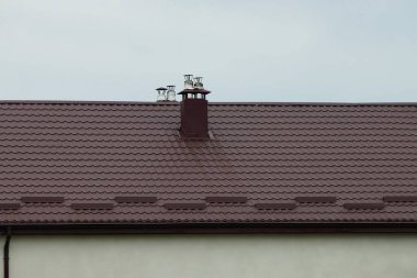 Bir evin kahverengi kiremitli çatısında gri gökyüzüne karşı metal bir baca.