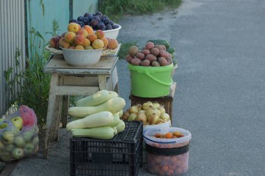 Sebzeler ve meyve kaseleri bir sandalyede ve kutular gri asfalt üzerinde duruyor.