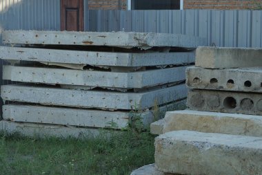 Metal bir çitin yanında yeşil çimlerde gri beton levhalar ve bloklar var.