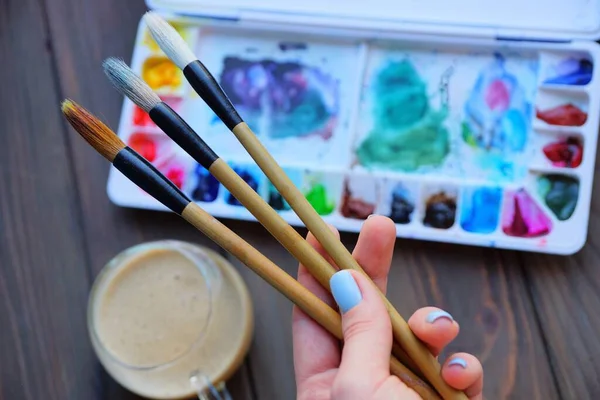 three brushes in the artist\'s hand on the background of a box with colored paints and a glass cup with brown coffee on the table