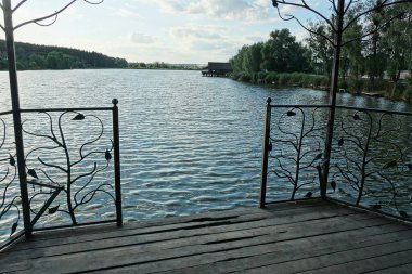 Kahverengi demir çitli, işlenmiş demir desenli ve göl suyunun arka planına karşı açık girişi olan bir taraçanın parçası.