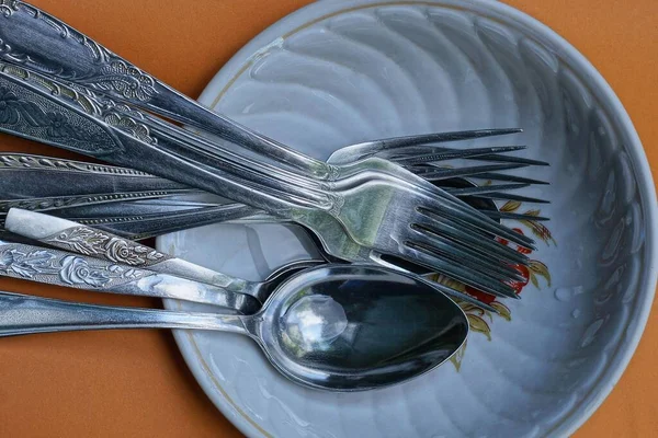 gray metal forks and spoons lie in a white wet plate on a brown table