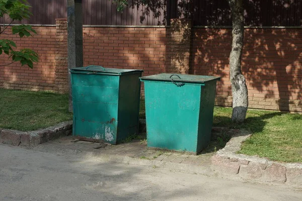 two iron green garbage cans stand in green grass by a gray asphalt road in front of a brown brick wall of a fence