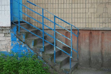Eski merdivenler gri beton basamaklar ve mavi demir parmaklıklar dışarıda kahverengi bir duvara çarpıyor.