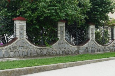 Gri taşlardan, tuğlalardan ve siyah demirden yapılmış uzun bir duvar çiti. Yeşil bitki örtüsü ve çimenlikte dövülmüş çubuklar.