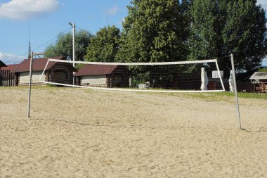 Beyaz voleybol ağı gri direkler üzerinde kahverengi kumlar üzerinde bir parkta mavi gökyüzü ve yeşil ağaçların arka planına karşı