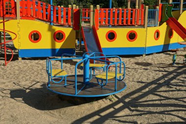 Mavi yuvarlak bir atlıkarınca, gri kumların üzerinde, bir sokak parkındaki renkli bir yapının yanında duruyor.