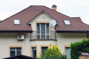 Gökyüzüne karşı kahverengi kiremitli çatı altında pencereleri olan özel bir evin ön cephesinde açık demir bir balkon.
