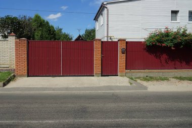 Kapalı bir kırmızı metal kapı ve caddede gri bir asfalt yolun kenarındaki çitin bir kısmı.