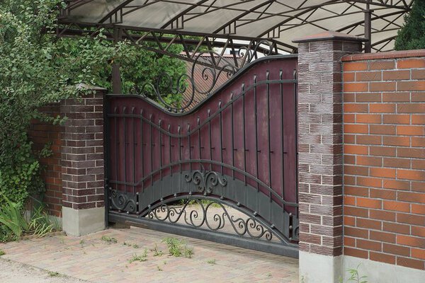 one metal gate with a black iron pattern and part of a wall of a fence made of brown bricks on the street