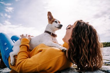 Güzel küçük bir köpek sahibinin üzerine oturmuş yüzüne bakıyor. Kadın Madrid 'de güneşli bir günde parkta uzanıyor. Aile köpeği açık hava yaşam tarzı