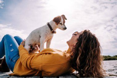 Güzel küçük bir köpek sahibinin üzerine oturmuş yüzüne bakıyor. Kadın Madrid 'de güneşli bir günde parkta uzanıyor. Aile köpeği açık hava yaşam tarzı
