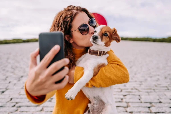 かわいい犬と一緒に公園にいる美しい女性 ペットとキスしながら携帯電話で自画像撮影中 ペットとテクノロジーライフスタイル — ストック写真