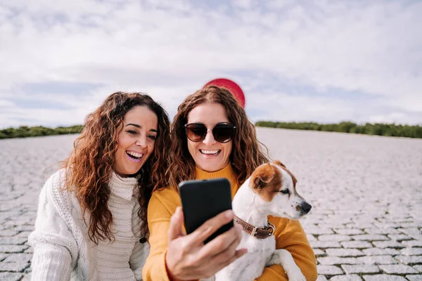 マドリードの公園で小さな犬と一緒に座っている2人の美しい女性が電話で写真を撮ろうとしています 犬とのテクノロジーライフスタイル — ストック写真