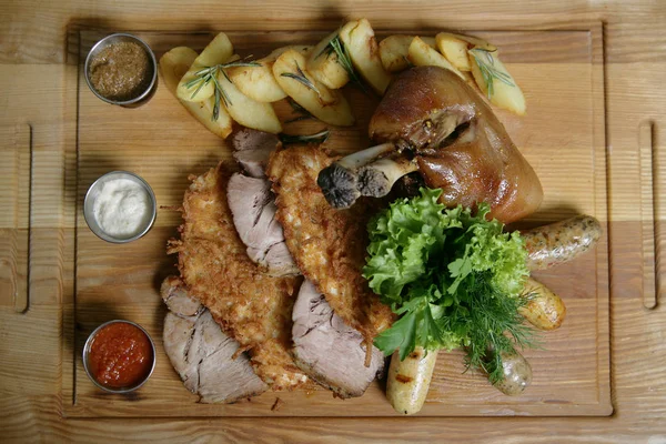 Mix food for a big company. A big delicious whole baked knuckle, fried meat chops, ham, potatoes. With greens and sauces. On a wooden board.