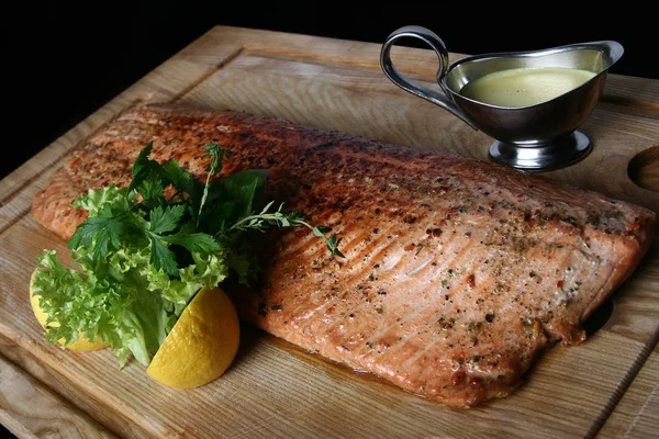 Grilled red fish. A large piece of delicious cooked fish. With greens, lemon and sauce on a wooden board close-up on a black background