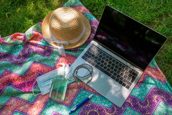 Laptop, face mask and hand alcohol gel sanitizer on blanket on a green grass lawn in the park or garden. Smart working from city park in a bright sunshine summer day.