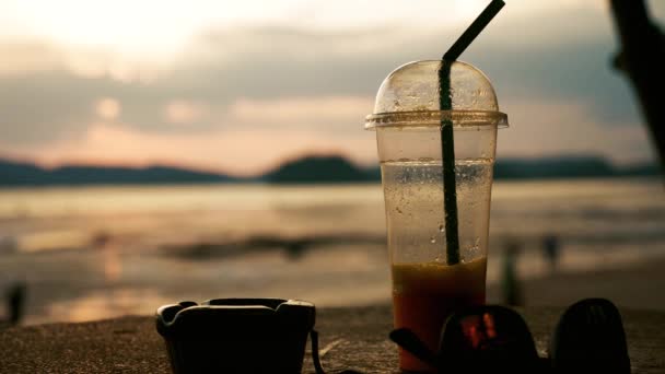 Šálek čerstvé šťávy na stole poblíž pláže při západu slunce — Stock video