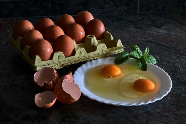 Two broken eggs on a plate, with mint leaves, along with half a dozen whole eggs