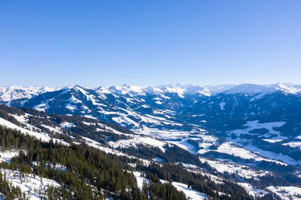 Vista desde un dron sobre montañas de invierno cubiertas de bosques y nieve en Alpes y pequeños pueblos —  Fotos de Stock