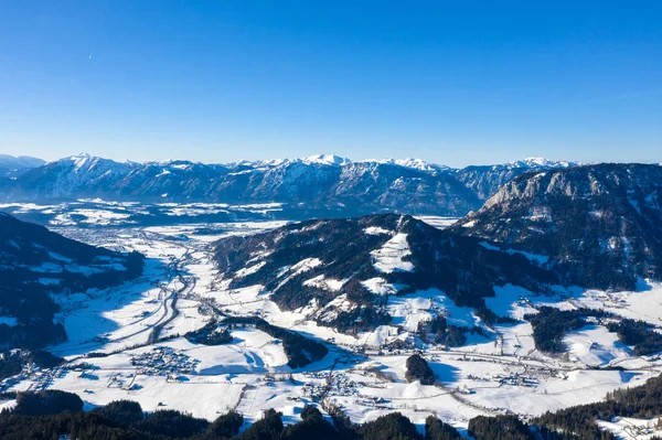 Vista desde un dron sobre montañas de invierno cubiertas de bosques y nieve en Alpes y pequeños pueblos —  Fotos de Stock