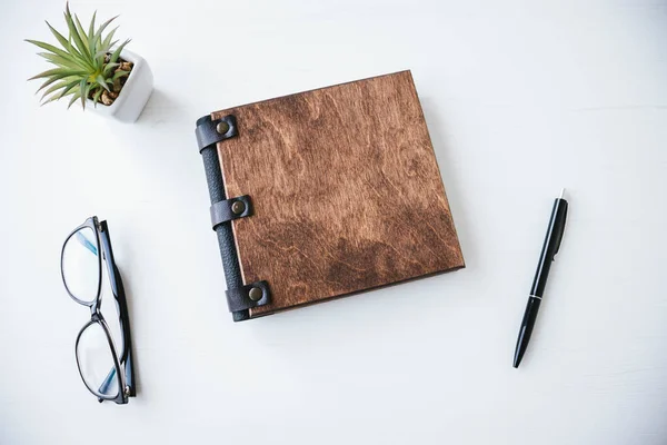 Ahşap kapaklı, kalemli, gözlüklü ve beyaz ahşap masadaki dekoratif bitkilerle dolu bir kitap..