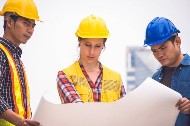 İnşaat işçileri ve mühendis ekibi koruyucu kask giyen ve proje detayları tartışmak ve planları ile yerinde duran, denetim için araçlar onları iş.