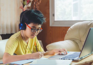 Kulaklık takan çocuk internette sohbet eden Skype öğretmeniyle masada oturuyor sınava hazırlanıyor ve ödevi için dizüstü bilgisayar kullanıyor ve online eğitim için E-öğrenim görüyor. çocuk konsepti öğrenmek.