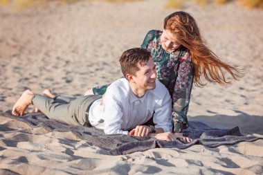 Genç çift sahilde kumda kıyafetlerle rahatlar, konuşur ve güler.